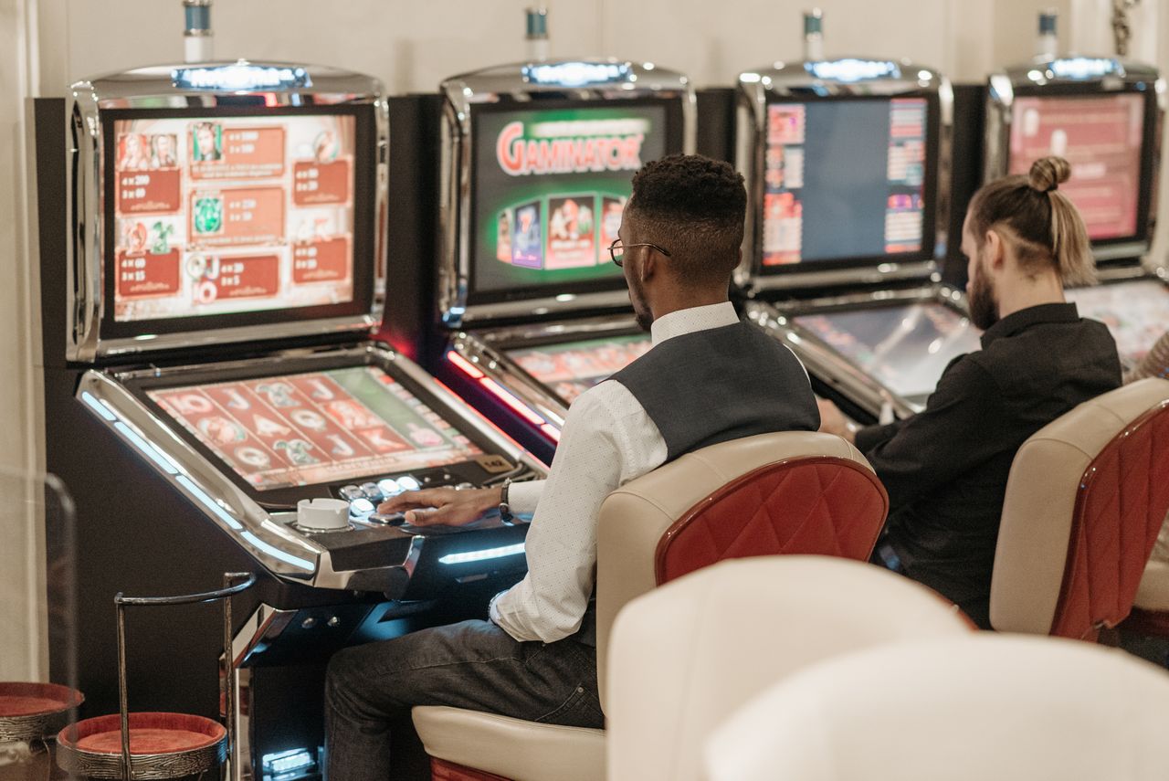 services-01 Two men engaged in gaming at a casino, seated at slot machines indoors.