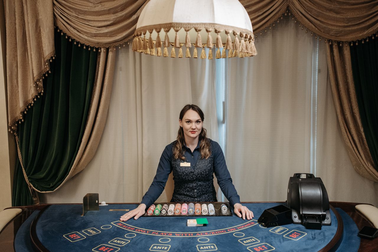gallery-6 A smiling female casino dealer at a gaming table surrounded by chips and cards indoors.